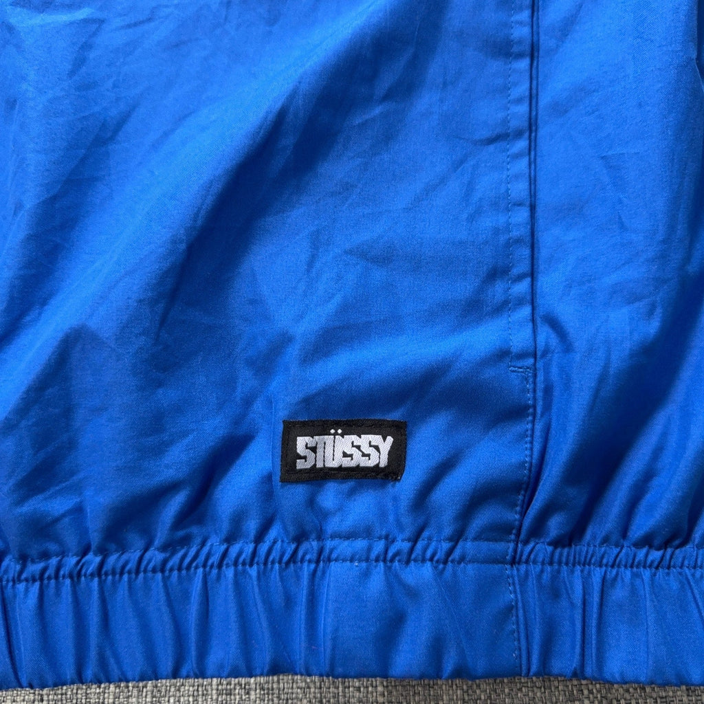Second Half Supply Co, LLC blue Stussy windbreaker jacket featuring a woven brand label on the elastic hem
